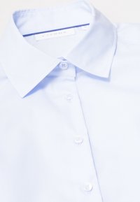 Chemise boutonnée bleu clair avec col classique, tissu lisse et boutons blancs. Étiquette visible près du cou.