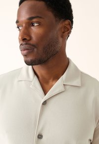 Ljusgrå skjorta med korta ärmar i texturerat tyg, med märkbar krage och två grå knappar synliga på framsidan.