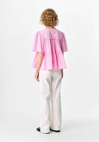 Top peplum rosa con maniche corte a sbuffo, corpetto arriccicato e bordi scallop, abbinato a jeans bianchi a gamba larga e scarpe nere.