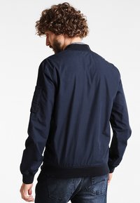 Marinfärgad bomberjacka i lätt tyg, med ribbad krage och ärmslut, sidofickor och en lapptäcksficka på vänster ärm.