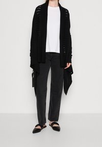 Svart stickad cardigan med öppen front, texturerade mönster, kombinerad med en vit t-shirt och svarta högmidjade byxor. Svarta platta skor.