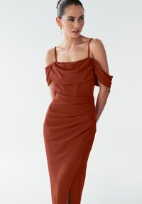 Nais, kes kannab pruuni off-shoulder kleiti õhukeste rihmadega, seisab ühetoonilise heleda tausta ees, kandes tilgakujulisi kuldseid ja pärlīt kaelakeed.