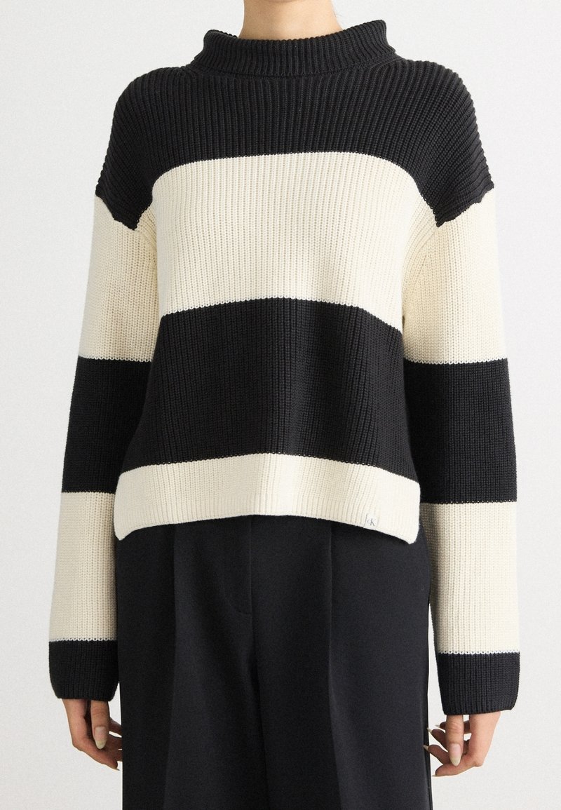 Femme portant un pull en tricot rayé horizontal noir et blanc à manches longues et un pantalon noir sur un fond uni.