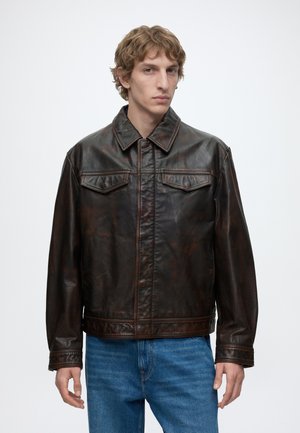 Junger Mann mit lockigem Haar, der eine dunkelbraune Lederjacke und blaue Jeans trägt, vor einem einfarbigen weißen Hintergrund stehend.