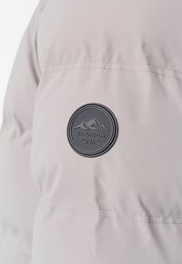 Jachetă puf gri deschis, cu un patch din cauciuc ce prezintă un design cu munți, textură netedă și detalii de cusătură subtile.
