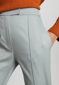 Svetle šedé tailored kalhoty s texturovanou tkaninou, se boční kapsou a hladkým povrchem. Spárované s oranžovým pleteným svetrem.