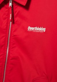 Rdeča jakna s srebrnim zadrgo in izvezenim belega besedilom na prsih, ki se glasi "Preveč razmišljam O VSEM od nekdaj".