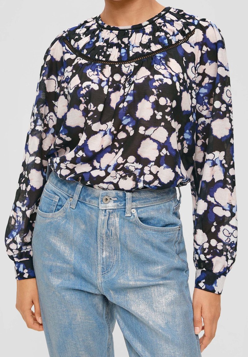 Schwarzbluse mit floralen Mustern in Blau- und Rosatönen, mit gerafftem Ausschnitt und langen Ärmeln, kombiniert mit hellblauen Jeans.