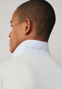 Světle šedý svetr s texturovanou tkaninou, s bílým límcem vyšitým jemným tónem "HACKETT", zobrazený ze zadní strany.