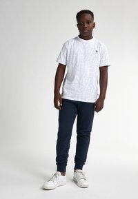 Camiseta blanca de manga corta con un patrón geométrico repetido, combinada con pantalones deportivos azul marino y zapatillas blancas con cordones.