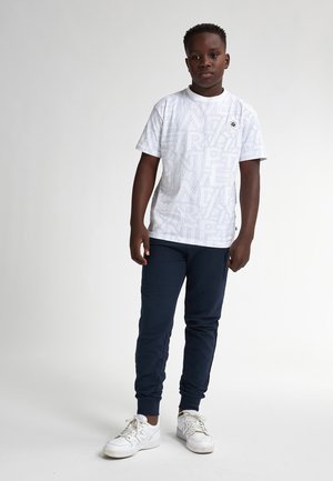 Camiseta blanca de manga corta con un patrón geométrico repetido, combinada con pantalones deportivos azul marino y zapatillas blancas con cordones.