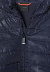 Tamno plavi pufasti kaput s teksturiranim materijalom, horizontalnim ukrasnim šavovima i kontrastnom narančastom unutrašnjom oznakom. Ima zatvaranje na rajsferšlus.