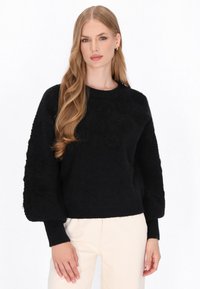 Czarny sweter z dzianiny z teksturowanym wzorem kwiatowym na przodzie oraz szerokimi rękawami. Ribbingowe mankiety i dolna krawędź. W połączeniu z jasnymi spodniami.