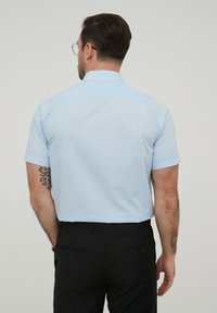 Chemise bleu clair à manches courtes avec un col à boutons, portée avec un pantalon noir. Tatouage visible sur le bras gauche. Texture de tissu lisse.