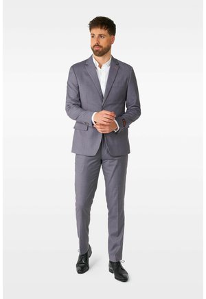 Grå skræddersyet jakkesæt med spids klap, slim fit og subtilt ternet mønster. Båret med en hvid skjorte og sorte sko.