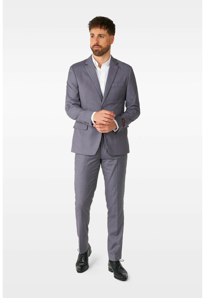 Grau maßgeschneiderter Anzug mit Reverskragen, slim fit und subtilen Karomuster. Kombiniert mit einem weißen Hemd und schwarzen Schuhen.