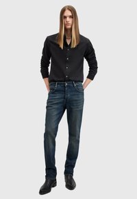 Camisa negra de botones con mangas largas, jeans de mezclilla azul y botas negras de tobillo; modelo con el cabello largo y lacio, de pie con las manos en los bolsillos.