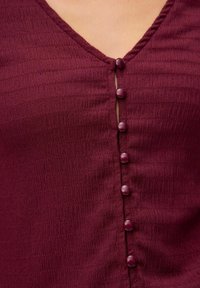 Dunkelroter, strukturierten Stoffoberteil mit V-Ausschnitt und sechs passenden Knöpfen an der Vorderseite. Der Stoff hat ein leicht geripptes Muster.