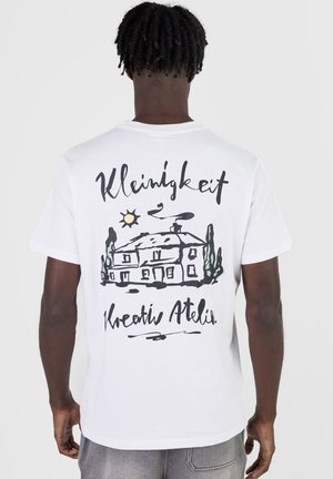 Mann mit dunkler Haut und lockigem Haar trägt weißes T-Shirt mit schwarzer Skizze eines Hauses, der Sonne, Bäumen und kursivem Text auf dem Rücken.