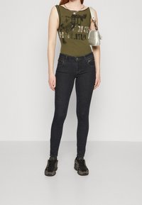 Jersey de cuerpo acanalado verde oliva con estampado gráfico, combinado con unos jeans ajustados oscuros. Zapatillas negras y grises, pequeña bolsa gris clara con correa de cadena.