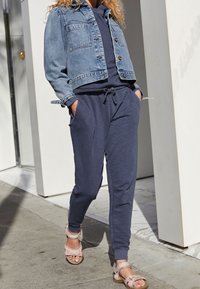 Denim-takki napitus edessä, kaksi rintataskua; tummansiniset jogger-housut, joissa on joustava vyötärö, yhdistettynä vaaleanpunaisiin remmissandaaleihin.