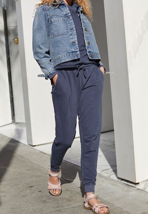 Giacca in denim con chiusura a bottoni, due tasche sul petto; pantaloni jogger blu navy con vita elastica, abbinati a sandali con cinturini rosa chiaro.