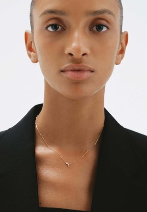 Primer plano de una persona que lleva un delicado collar de oro con tres pequeños colgantes de piedras preciosas, vestida con un blazer negro.