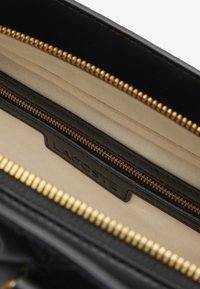 Interno di una borsa in pelle nera con zip dorata e fodera in tessuto beige. Presenta un'etichetta in pelle impressa con "Lacoste."