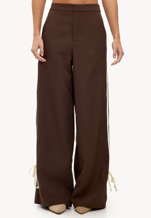 Pantalons larges marron avec des rayures blanches sur les côtés et des liens près des chevilles, portés avec des chaussures beige à bout pointu, présentés de la taille aux pieds.