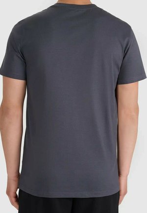 Osoba ubrana w gładki ciemnoszary t-shirt z krótkim rękawem, widziana od tyłu, stojąca na tle jednolitego jasnego tła.