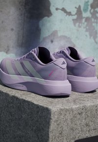 Para jasnofioletowych butów do biegania Adidas z białymi paskami, umieszczona na kamiennym parapecie na tle teksturowanej ściany.