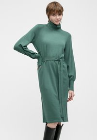 Strikkekjole i grønn med lange ermer og høyt hals. Har belte i midjen og rekker til midten av leggen. Glatt tekstur, minimalistisk design.