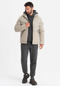 Giacca imbottita beige con cappuccio, caratterizzata da un design trapuntato, cerniere frontali e pantaloni sportivi grigi. Scarpe sportive scure completano l'outfit.