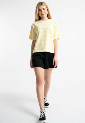 Młoda kobieta idzie do przodu, ubrana w żółty koszulkę w paski, czarne szorty i czarno-białe tenisówki na białym tle.