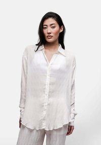 Camicia bianca con bottoni, dotata di pieghe verticali, maniche lunghe e colletto. Presenta una leggera lucentezza e un orlo curvato.