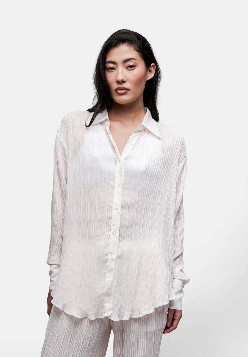 Camicia bianca con bottoni, dotata di pieghe verticali, maniche lunghe e colletto. Presenta una leggera lucentezza e un orlo curvato.
