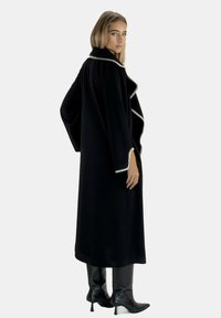 Zwarte lange jas met een sjaalkraag, witte bies langs de randen en brede mouwen. Model draagt zwarte kniehoge laarzen en staat zijwaarts.