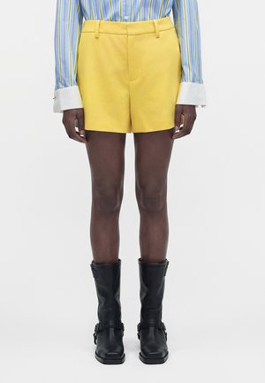 Personne portant un short jaune sur mesure, une chemise bleue rayée avec des poignets blancs et des bottes en cuir noires mi-mollet, debout, droite.