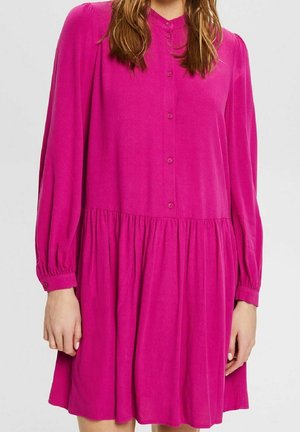 Kvinde iført en skrigende pink langærmet kjole med knapper, med en samlet talje og løs nederdel mod en ensfarvet baggrund.