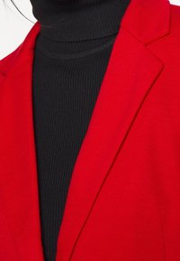 Red blazer with a notched collar, textured fabric, paired with a black ribbed turtleneck. Closely cropped to focus on the neckline and collar.