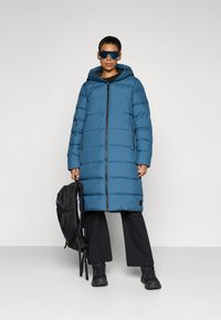 Long puffer coat in blue, featuring a zip front, hood, and quilted design. Paired with black pants and a black backpack.