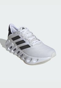 Scarpa sportiva bianca con tomaia testurizzata, accenti neri, tre strisce nere distintive e un design del suola con un sistema di ammortizzazione unico.