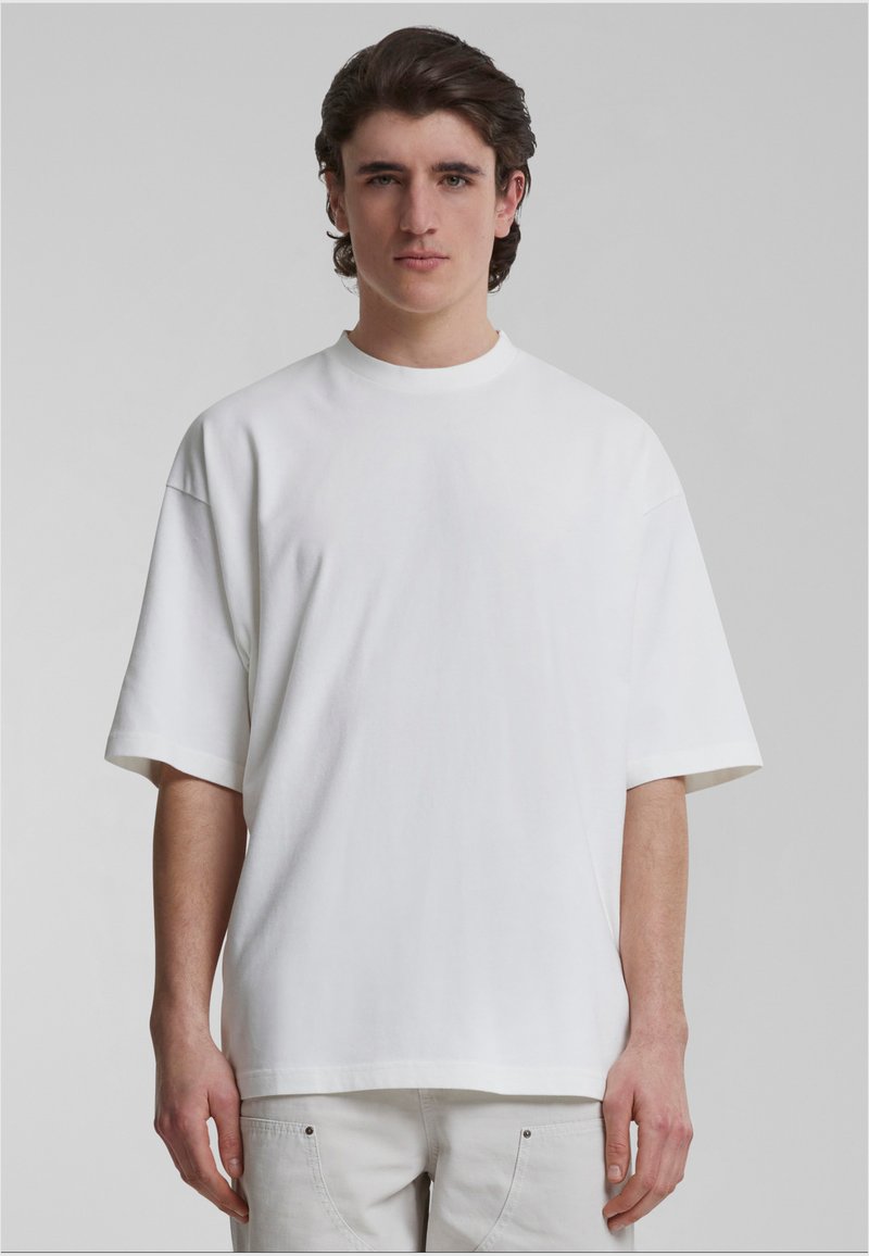 Weißes Baumwoll-T-Shirt mit kurzen Ärmeln und rundem Ausschnitt. Hat eine lockere Passform und eine glatte Textur, getragen mit hellfarbenen Hosen.