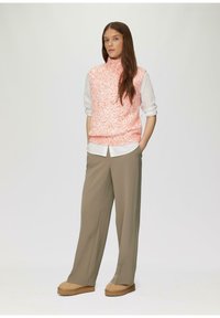Licht koraalkleurig gebreid mouwloos topje met een hoge kraag, gelaagd over een witte blouse. Gecombineerd met losse beige broek en tan platform schoenen.