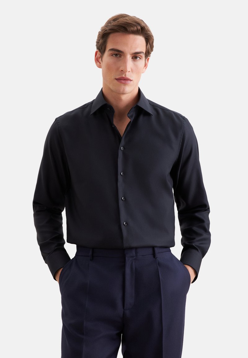 Jeune homme aux cheveux châtain clair portant une chemise boutonnée bleu marine foncé et un pantalon assorti, debout les mains dans les poches devant un fond blanc.