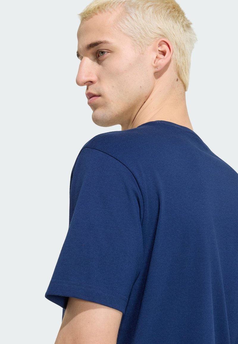 Jeune homme aux cheveux courts décolorés portant un t-shirt bleu foncé uni, tourné vers la gauche et regardant par-dessus son épaule sur fond blanc.