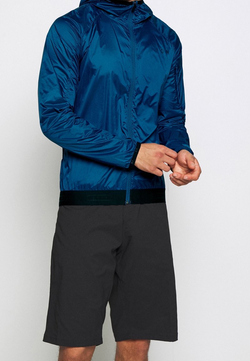 Man i en blå lättviktsjacka med dragkedja och huva, samt svarta knä-långa shorts, justerar ärmvesken mot en vit bakgrund.