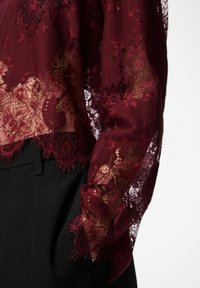 Caursrdzama burgundijas mežģīņu blūzes piedurkne ar ziedu rakstiem pār melnām biksēm, roka daļēji ielikta kabatā, uz vienveidīga fona.