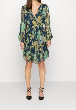 Femme portant une robe fleurie bleu marine transparente avec de longues manches bouffantes et des chaussures noires à talons hauts à brides, debout devant un fond uni.