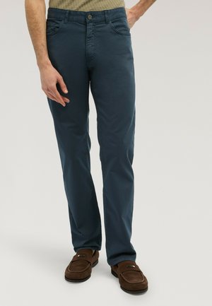 Männliches Model trägt dunkelblaue, gerade geschnittene Hose und braune Wildleder-Loafer vor hellgrauem Hintergrund.